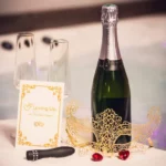 Image d'une table dressée devant un jacuzzi, avec une bouteille de champagne fermée et un masque en dentelle doré posé dessus. À côté, deux coupes de champagne vides et une carte blanche avec le logo Loving Up. Devant la carte, un vibromasseur noir orné de strass rose et doré est placé à côté de deux pierres rouges.