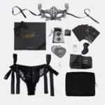 La Love Box Shhh... est présentée avec tous ses éléments. À gauche, en haut de la boîte, se trouve un masque noir en dentelle avec deux ficelles. En bas de la boîte, une culotte noire en dentelle s'attache sur les côtés avec des rubans noirs. À droite de l'image, on trouve de haut en bas un jeu de 2 dés, un jeu de cartes spicy disposé en forme d'éventail, un jeu de gages, un cockring vibrant extensible, un stimulateur clitoridien rechargeable, un bandeau-lien en satin noir extensible et une trousse noire avec son charm doré.