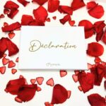 Image du coffret blanc avec le nom "Déclaration" écrit en doré. Entourée de pétales de rose rouge et de petits strass pailletés en forme de cœur.